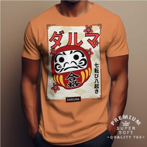 Peut inclure: T-shirt orange avec un motif de poupée Daruma. Le graphisme comprend du texte japonais et une poupée Daruma rouge et blanche avec un visage détaillé. Le t-shirt porte également l'inscription "DARUMA" et "PREMIUM SUPER SOFT QUALITY TEE."