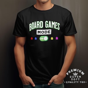Può includere: T-shirt nera con le parole "BOARD GAMES MODE" in verde e bianco. La parola "MODE" è sopra un pulsante verde "ON" con icone di pedine. La maglietta ha anche il testo "PREMIUM SUPER SOFT QUALITY TEE".