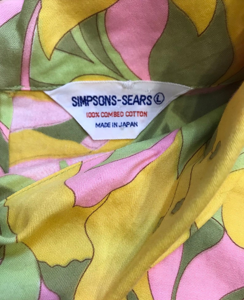 K&ouml;nnte beinhalten: Ein Hemd mit gelbem, rosa und gr&uuml;nem Blumendruck und einem wei&szlig;en Etikett mit der Aufschrift "SIMPSONS-SEARS&reg; 100% GEK&Auml;MMTE BAUMWOLLE HERGESTELLT IN JAPAN".