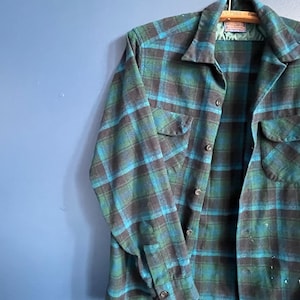 Puede incluir: Una camisa de cuadros vintage en tonos turquesa, verde y marrón cuelga de una percha de madera. La camisa tiene una parte delantera abotonada, dos bolsillos en el pecho y puños abotonados. La tela tiene un aspecto texturizado.