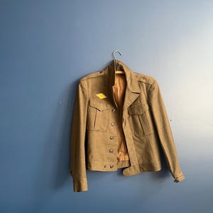 May include: Olive green jacket with four front pockets and button fastenings. The jacket is hanging on a wooden hanger against a blue wall. A yellow tag is attached.