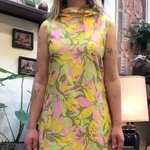 K&ouml;nnte beinhalten: Ein &auml;rmelloses Kleid mit hohem Kragen und einem Blumendruck in Gelb-, Rosa- und Gr&uuml;nt&ouml;nen.