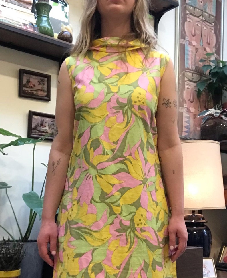K&ouml;nnte beinhalten: Ein &auml;rmelloses Kleid mit hohem Kragen und einem Blumendruck in Gelb-, Rosa- und Gr&uuml;nt&ouml;nen.