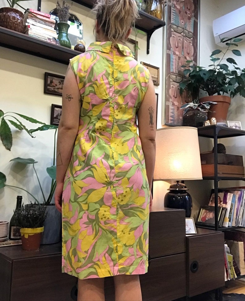 K&ouml;nnte beinhalten: Ein &auml;rmelloses Kleid mit einem pinkfarbenen, gelben und gr&uuml;nen Blumendruck. Das Kleid hat einen Kragen und einen Knopfverschluss auf der R&uuml;ckseite.