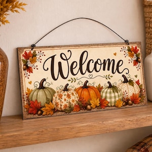 May include: A rectangular hanging sign with a cream-colored background featuring the word Welcome in dark brown script. The design includes five pumpkins in various colors and patterns, surrounded by autumn leaves, acorns, and small hearts.