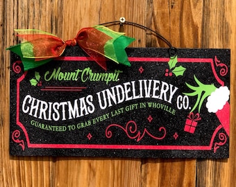 May include: A black and green Christmas sign with the text "Mount Crumpit Christmas Undelivery Co. Guaranteed to grab every last gift in Whoville." The sign has a red border and a green and red bow.