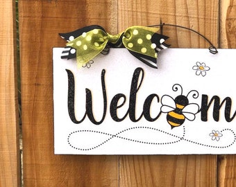 May include: White wooden welcome sign with black glitter lettering and a bee illustration. The sign is decorated with a green and black polka dot bow.