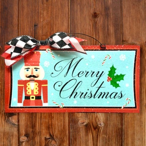 May include: A red and white Christmas sign with a nutcracker illustration and the text "Merry Christmas". The sign has a red border and a blue background with white snowflakes and candy canes.