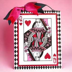 May include: A decorative Queen of Hearts playing card design. The card features a red, black, and white color scheme with a glittery border and a pink bow at the top. The card is a rectangular shape.