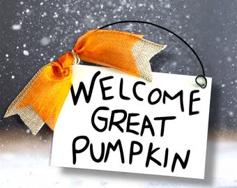 May include: A white rectangular sign with the words "WELCOME GREAT PUMPKIN" in black lettering. The sign is adorned with an orange ribbon with gold trim and a black string for hanging. The background is a snowy scene.