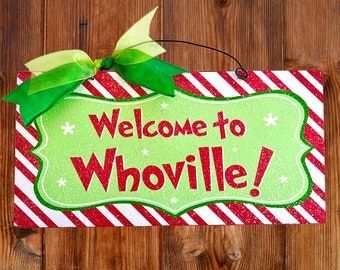 May include: A red and white striped sign with a green welcome sign that says "Welcome to Whoville!" with snowflakes on the sign. The sign is decorated with a green bow.