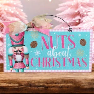 May include: A rectangular Christmas sign with a pink and teal color scheme. The sign features a nutcracker illustration and the phrase "Nuts about Christmas" in pink lettering. A pink bow and wire hanger are attached.