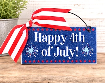 May include: A rectangular blue sign with the text "Happy 4th of July!" in white. The sign is decorated with red stars and blue and white fireworks. A red and white striped ribbon is attached to the top.