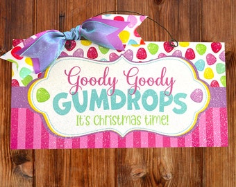 May include: A rectangular sign with the words "Goody Goody Gumdrops" and "It's christmas time!" in a decorative font. The sign has a pink and white striped background and is adorned with a colorful bow.