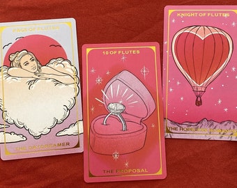 May include: Three tarot cards with pink and gold backgrounds. The first card is the Page of Flutes and features a woman laying in a cloud with the text "The Daydreamer". The second card is the 10 of Flutes and features a ring in a heart-shaped box with the text "The Proposal". The third card is the Knight of Flutes and features a hot air balloon in the shape of a heart with the text "The Hopeless Romantic".