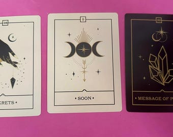 May include: Three tarot cards with gold and black designs on a pink background. The first card has a black hand with a gold bracelet holding a pendulum. The text on the card reads "Secrets". The second card has a gold triple moon symbol with a gold arrow pointing up. The text on the card reads "Soon". The third card has a gold crystal on a black background. The text on the card reads "Message of Peace".