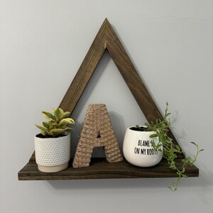 May include: A triangular wooden shelf with a brown stain. The shelf has a small potted plant, a woven letter A, and a white pot with the text "Blame it on my roots".