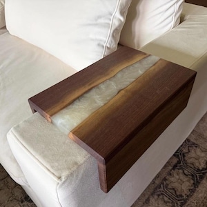 May include: A rectangular sofa arm tray constructed from two dark brown walnut wood slabs joined by a central strip of translucent, pearlescent white epoxy resin. The tray features a flat top surface and two side panels that drape over the arm.