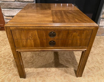 May include: A wooden end table with two drawers and a square top. The drawers have brass pulls with a circular design. The table has a dark brown finish.
