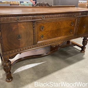 May include: A vintage wooden buffet with intricate carvings and ornate hardware. The buffet has four drawers and two doors, and it is supported by four turned legs. The buffet is finished in a rich brown color.