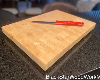 May include: A light-colored, rectangular butcher block cutting board with a red-handled knife resting on top. The cutting board is made of wood and has a patterned surface. The text "BlackStarWoodWorkMN" is in the bottom right corner.
