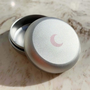 May include: A silver round tin with a pink crescent moon design on the lid. The tin is open and the inside is visible.