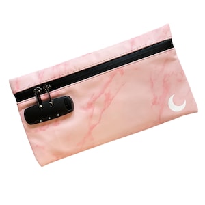 May include: A rectangular pouch featuring a light pink marble pattern, a black horizontal zipper closure, and a black three-digit combination lock. A small white crescent moon icon is printed on the lower right corner of the pink surface.