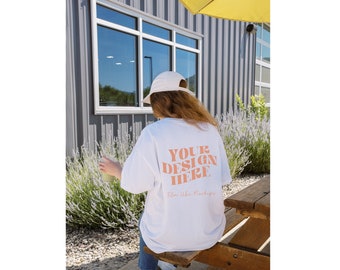 May include: White Gildan 5000 t-shirt mockup with the text "YOUR DESIGN HERE" printed on the back. The t-shirt is being worn by a person sitting on a wooden bench outdoors.