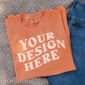 May include: An orange t-shirt with the text "YOUR DESIGN HERE" in white block letters. The shirt is folded and placed next to a pair of blue jeans on a patterned surface. The t-shirt has a crew neck.
