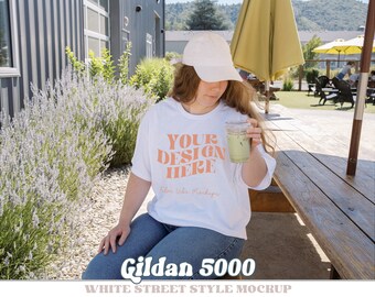 May include: White Gildan 5000 t-shirt mockup with the text "Your Design Here" in orange. A person is wearing the t-shirt and holding a glass of iced tea. The person is sitting at a wooden table outdoors.