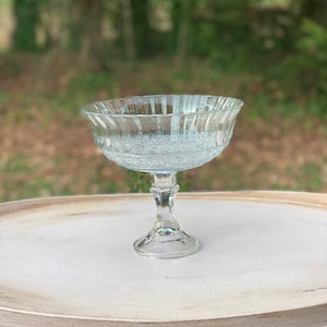 May include: A clear glass pedestal bowl with a fluted rim and a textured base. The bowl is sitting on a light brown table.
