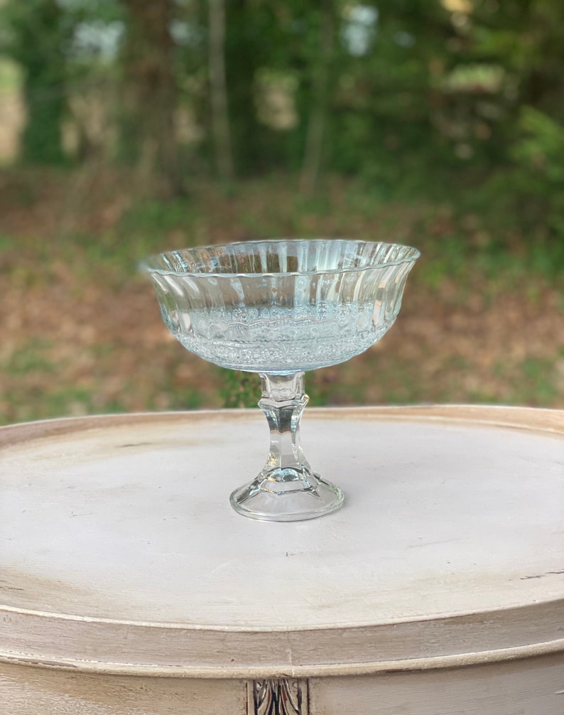 May include: A clear glass pedestal bowl with a fluted rim and a textured base. The bowl is sitting on a light brown table.