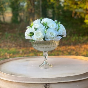 May include: A white floral arrangement in a clear glass vase with a decorative pattern. The vase has a pedestal base and is sitting on a white table.