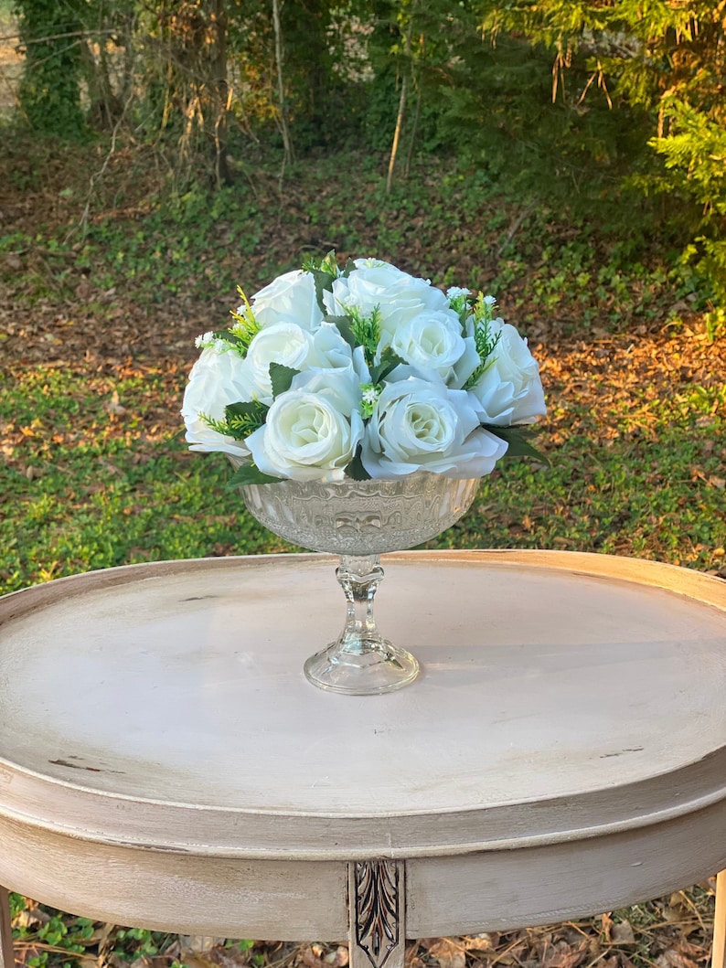 May include: A white floral arrangement in a clear glass vase with a decorative pattern. The vase is on a white table with a distressed finish.