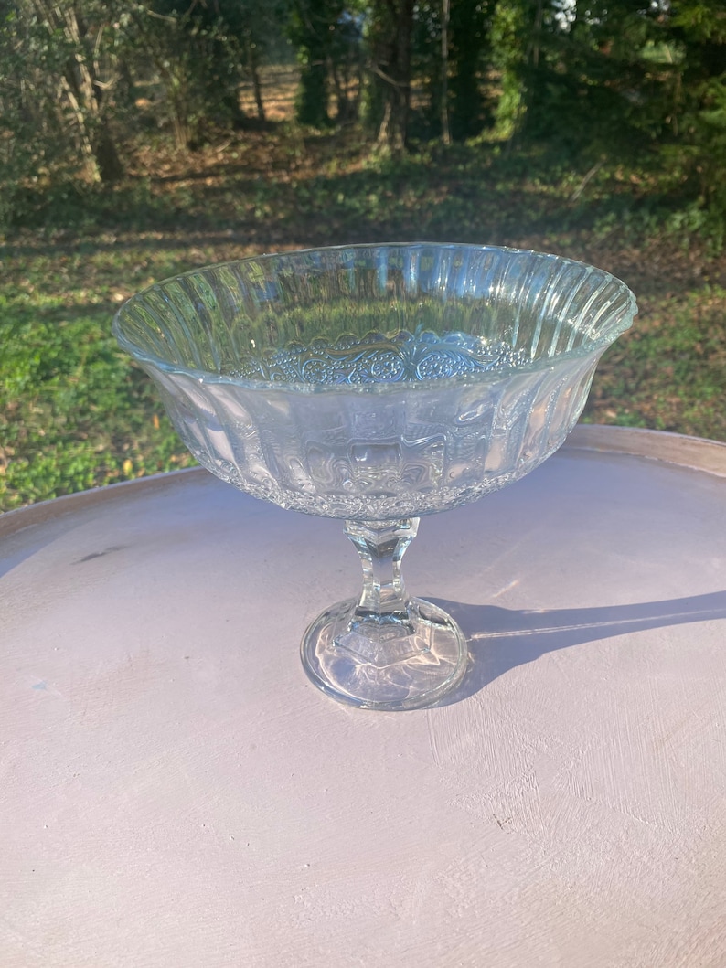 May include: A clear glass pedestal bowl with a fluted rim and a decorative pattern. The bowl is on a white surface.