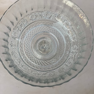 May include: A clear glass cake stand with a decorative pattern around the edge. The stand has a raised center and a scalloped edge.
