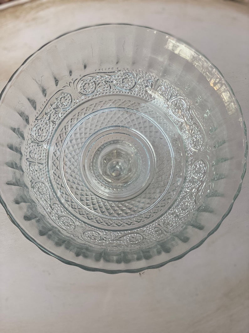 May include: A clear glass cake stand with a decorative pattern around the edge. The stand has a raised center and a scalloped edge.