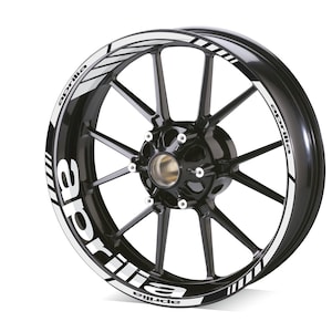 Può includere: Un cerchio di ruota di moto nero con una striscia bianca e la parola "Aprilia" ripetuta attorno al cerchio.