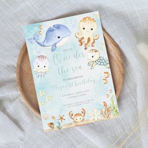 Puede incluir: Una tarjeta de invitación azul y blanca con una ilustración de acuarela de una ballena, un pulpo, una medusa, una tortuga y un cangrejo. El texto de la tarjeta dice: "Únete a nosotros Oneder the sea para celebrar el 1er cumpleaños de Aryas Sábado, 10 de marzo - 8:30am - 1:30pm Casa de la abuela y el abuelo 123 Anywhere Street"