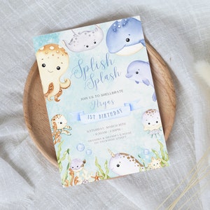 Puede incluir: Una tarjeta de invitación azul y blanca con una ilustración de acuarela de criaturas marinas, incluyendo un pulpo, una ballena, una raya, un delfín, una medusa y un narval. El texto en la tarjeta dice "Splish Splash Join us to shellbrate Aryas 1st Birthday Saturday March 10th 11:30am - 1:30pm Grandma & Grandpa's House 123 Anywhere Street."