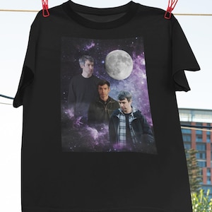 Pode incluir: Camiseta preta com um gráfico de três homens contra um fundo espacial, com uma lua cheia e uma nebulosa roxa e branca. A camiseta está pendurada em um varal com prendedores de roupa vermelhos.