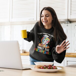 May include: A black sweatshirt with a colourful unicorn wearing a chef's hat and holding a whisk. The text on the sweatshirt reads "I just baked you some shut the f*cupcakes".