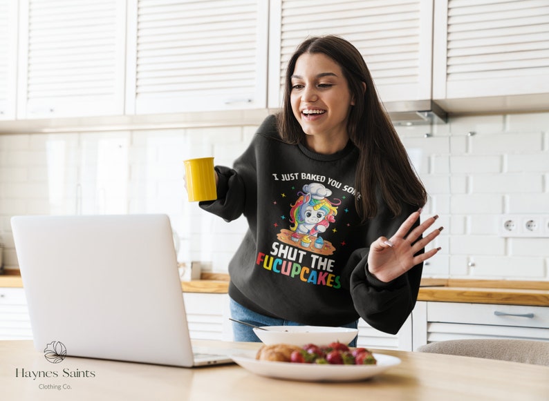 May include: A black sweatshirt with a colourful unicorn wearing a chef's hat and holding a whisk. The text on the sweatshirt reads "I just baked you some shut the f*cupcakes".
