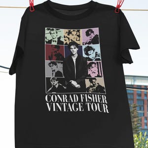 Könnte beinhalten: Schwarzes T-Shirt mit einer Collage von Bildern einer Person und dem Text "CONRAD FISHER VINTAGE TOUR" in Weiß. Das T-Shirt hängt an einer Wäscheleine mit roten Wäscheklammern. Das Markenlogo "Haynes Saints Clothing Co." ist sichtbar.