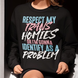 Może przedstawiać: Czarna bluza z napisem "RESPECT MY TRANS HOMIES OR I'M GONNA IDENTIFY AS A PROBLEM" w kolorze niebieskim, r&oacute;żowym i białym. Bluza pochodzi od Haynes Saints Clothing Co.