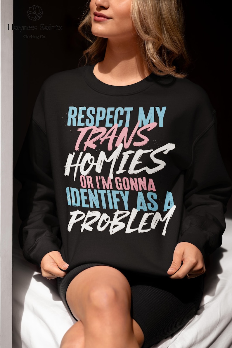 Może przedstawiać: Czarna bluza z napisem "RESPECT MY TRANS HOMIES OR I'M GONNA IDENTIFY AS A PROBLEM" w kolorze niebieskim, r&oacute;żowym i białym. Bluza pochodzi od Haynes Saints Clothing Co.