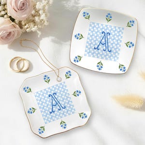 May include: Two white square dishes with gold trim, featuring a blue and white checkered pattern and the letter "A". Each dish is decorated with blue floral accents. A gold necklace and two gold rings are also present.