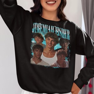 May include: Black sweatshirt featuring a graphic of the name "Jeremiah Fisher" in turquoise lettering, with multiple images of a young man. The sweatshirt has a crew neck and long sleeves. The brand name "Haynes Saints Clothing Co." is printed at the bottom.