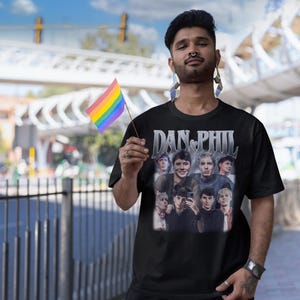 Może przedstawiać: Czarny t-shirt z grafiką przedstawiającą kilka os&oacute;b i napisem "DAN & PHIL". Osoba na zdjęciu trzyma tęczową flagę. Nazwa marki "Haynes Saints Clothing Co." jest widoczna w lewym g&oacute;rnym rogu.