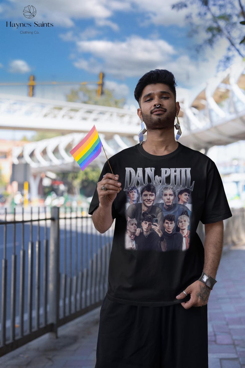 Może przedstawiać: Czarny t-shirt z grafiką przedstawiającą kilka os&oacute;b i napisem "DAN & PHIL". Osoba na zdjęciu trzyma tęczową flagę. Nazwa marki "Haynes Saints Clothing Co." jest widoczna w lewym g&oacute;rnym rogu.